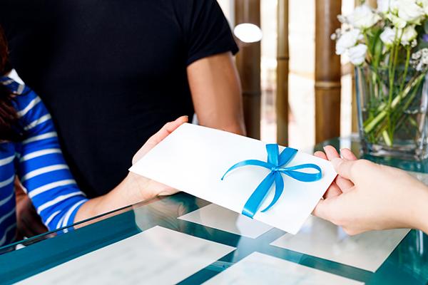 One person hands a white envelope with a blue ribbon to another person while a third person looks on in the background.